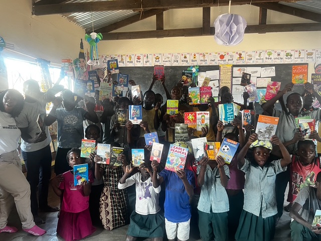 A group of children and adults standing inside a classroom, holding up various colorful books and smiling. The children are wearing school uniforms, while the adults are casually dressed. The classroom walls are decorated with educational posters and learning materials. The group is celebrating, with books held high, creating an atmosphere of joy and excitement about reading and learning.