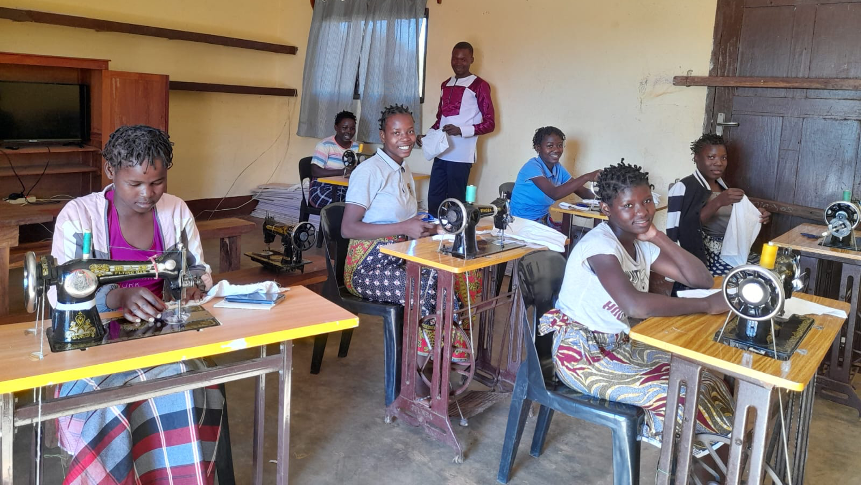 A group of young women sitting at sewing machines in a classroom setting, smiling and working on their sewing projects. The women are dressed in casual clothes, and the room is simply furnished with wooden tables and chairs. An instructor stands in the background, watching over the students. The atmosphere is focused and positive, conveying a sense of empowerment and skill-building.