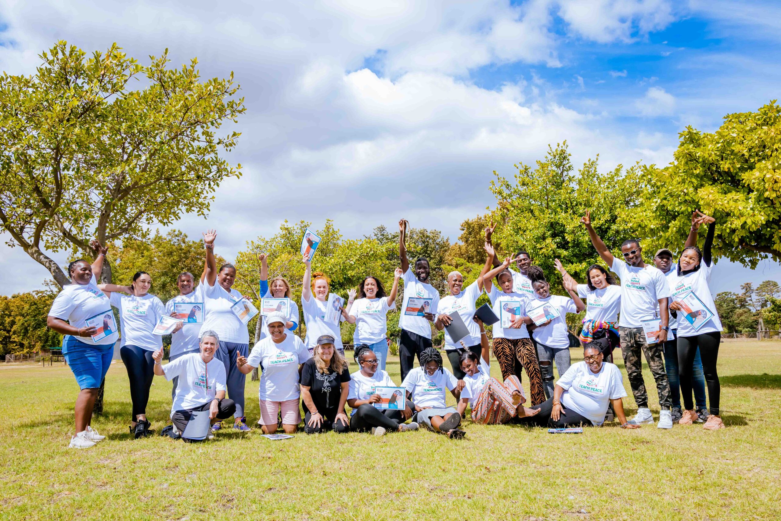 A group of people standing and kneeling outdoors in a grassy area, celebrating and smiling for the camera. They are wearing matching white T-shirts, and some are holding certificates or awards. The group appears diverse, with individuals of various ages and backgrounds. Green trees and a partly cloudy sky serve as the backdrop, creating a bright, cheerful atmosphere.